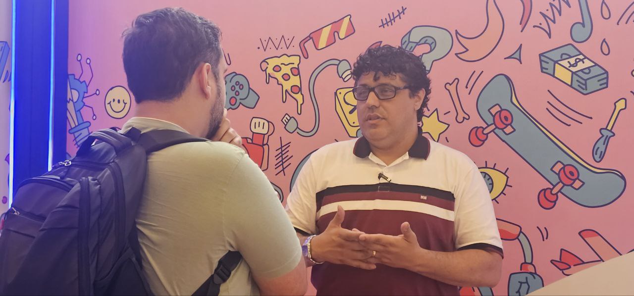 Jose JR conversando com uma conferencista que esta de costas na foto enquanto jose Jr esta de frente para ele, usando camiseta polo branco a roxo