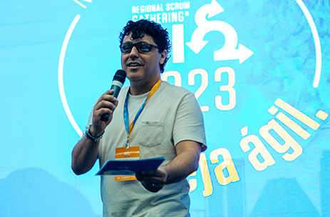 Jose JR esta segurando um microfone sendo Mestre de cerimonia . esta em pe no palco.. ao fundo esta a logo do SGRIO 2023