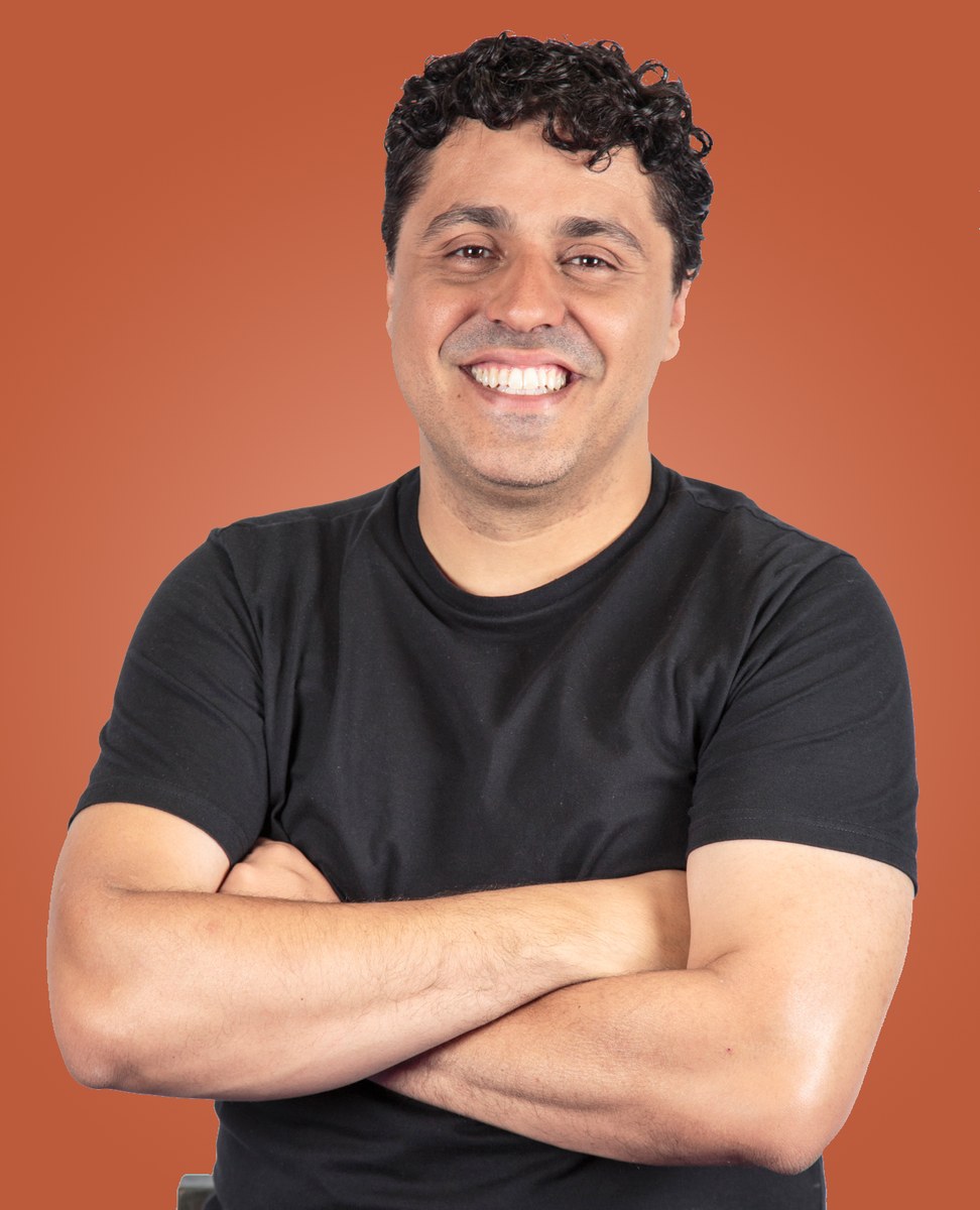 Jose JR de braços cruzados, camiseta preta, sorrindo, sobre fundo terracota